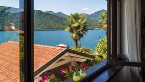 A view from a window overlooking a lake with mountains, a palm tree, and a tiled roof. - LA PLAYA: Villa with a Private Beach and All-Inclusive Sports  (Pettenasco)
