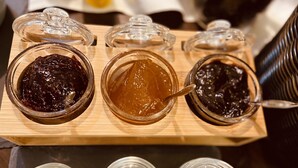 Una tabla de madera con seis frascos de vidrio que contienen diversos tipos de untables.