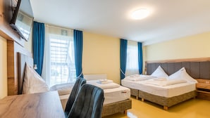 Habitación de hotel con dos camas individuales, una mesa de madera, un televisor montado en la pared y una ventana con cortinas.
