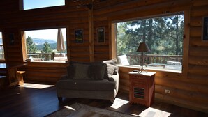 L’intérieur d’un chalet accueillant, avec un canapé, une table en bois et une fenêtre donnant sur une forêt et des montagnes.