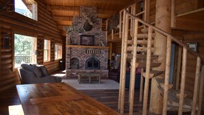 L’intérieur d’une cabane en rondins, avec une cheminée, un escalier en bois et un coin repas.