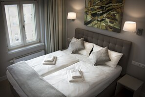 Cama doble con dos almohadas y dos toallas dobladas, una ventana con cortinas y dos lámparas fijadas a la pared.