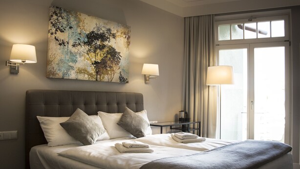 Ein Hotelzimmer mit einem großen Bett, zwei Wandlampen, einem Bild an der Wand und Blick ins Freie durch ein Fenster.