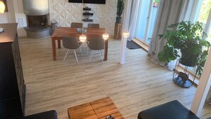 Ein modernes Wohnzimmer mit Esstisch aus Holz, bequemen Sitzgelegenheiten, einem flachbildfernseher an einer strukturierten Wand und einer großen Küstpflanze.
