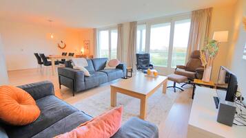 Een moderne woonkamer met een grijze bank, een salontafel en een bruine leren fauteuil. In de achtergrond is een eethoek met een tafel en stoelen te zien.