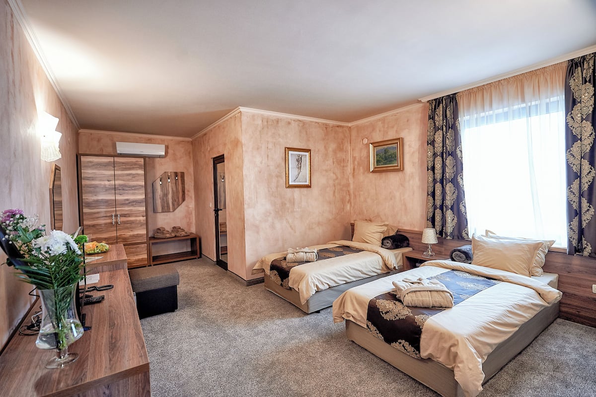 A hotel room with two beds, a desk with a vase of flowers, a wardrobe, and a large window with curtains.