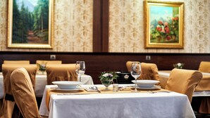 A dining area with tables set for two, each with a white tablecloth, silverware, and a glass of water. The walls are adorned with framed paintings. - Platinum Image Hotel (Elin Pelin)