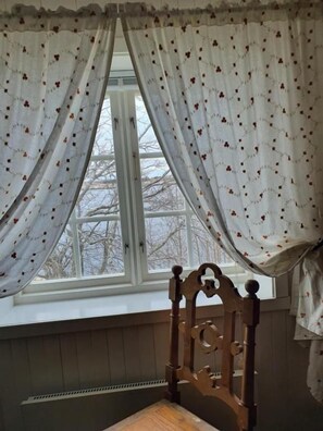 A room with a wooden chair, a window with white curtains, and a view of trees outside. - Handelsstedet Forvik (Vevelstad)