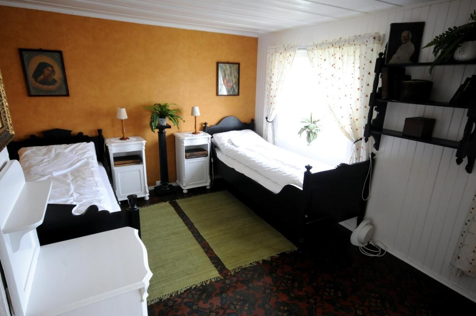 A room with two beds, a nightstand with a lamp, a small table with a plant, and a radiator.