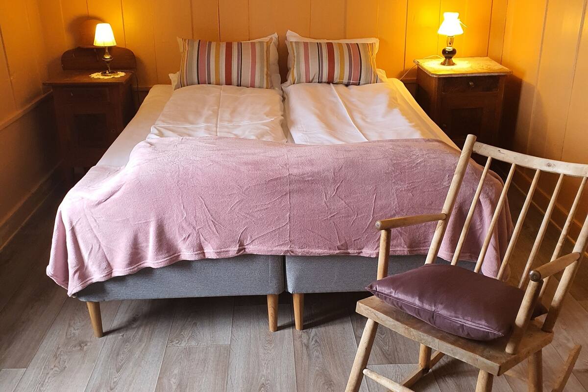 Un dormitorio con una cama, dos mesitas de noche, un sillón mecedora y dos cuadros en la pared.