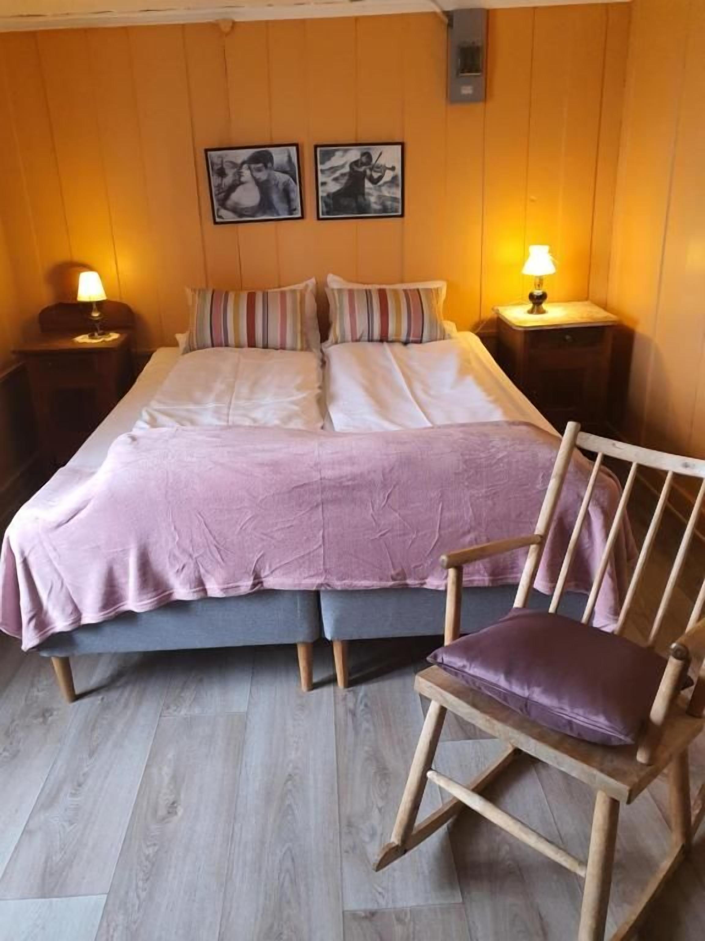 Une chambre à coucher comprenant un lit, deux tables de chevet, une chaise en bois et deux lampes fixées au mur.