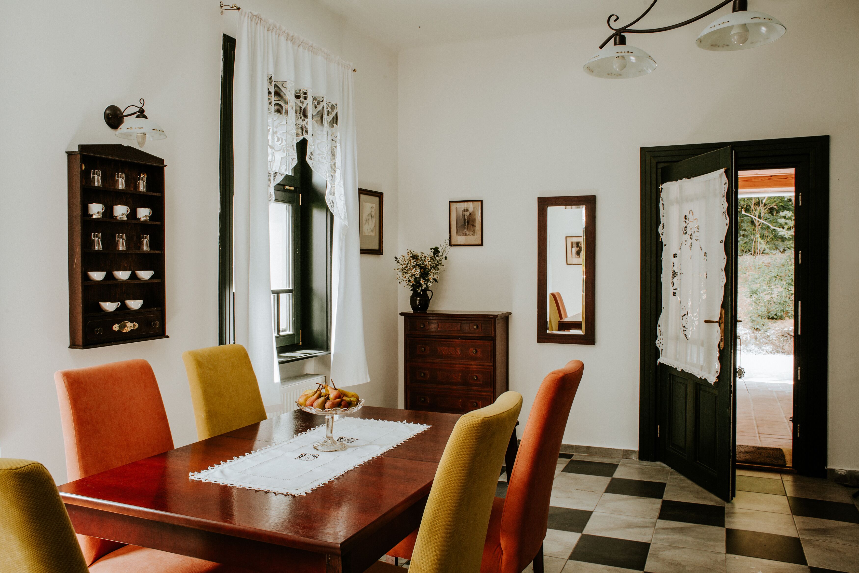 Comedor con una mesa de madera, sillas naranjas y amarillas, un aparador con vajilla y piso a cuadros.