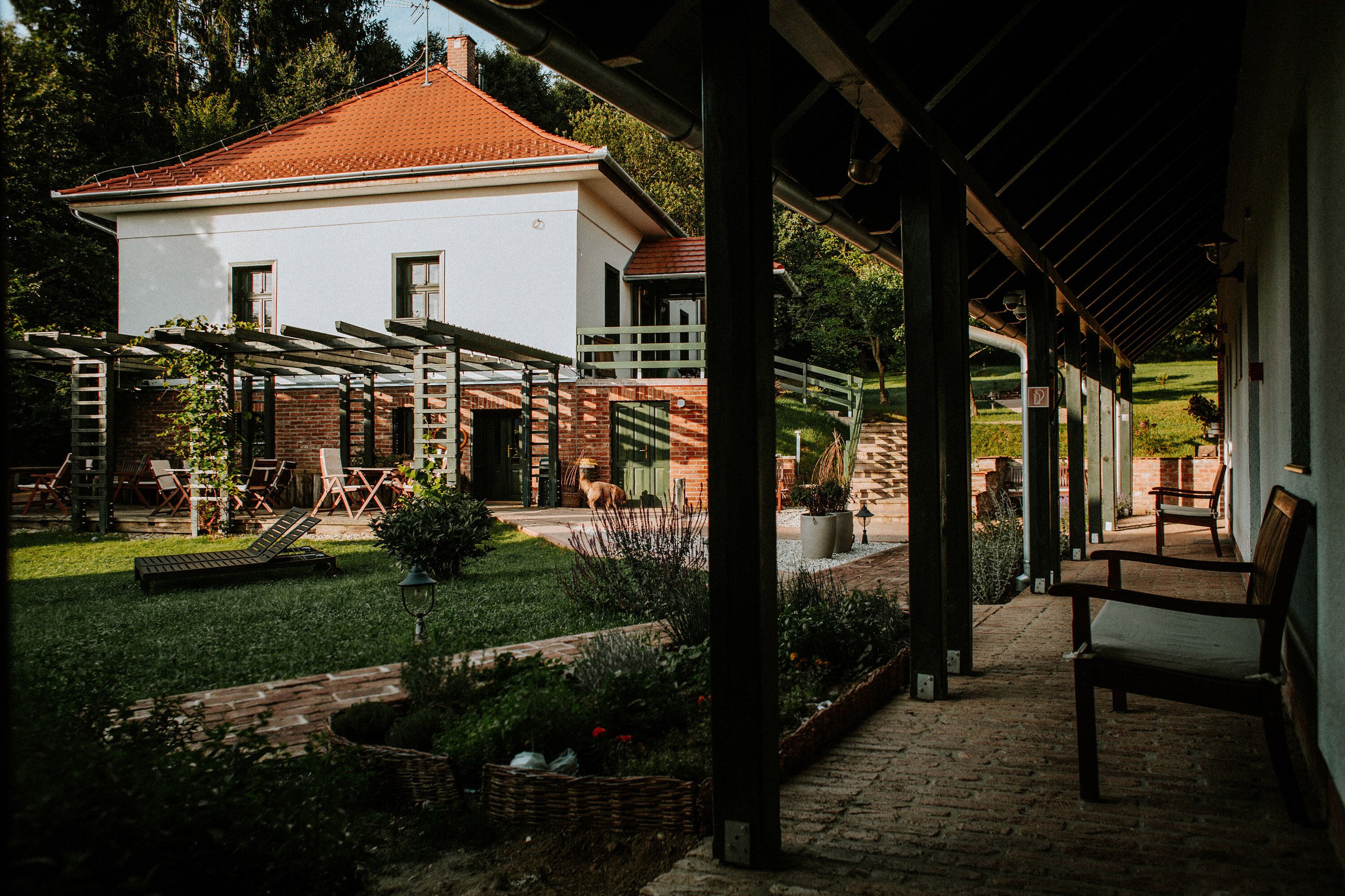 A covered outdoor area with a white building, a brick wall, and a grassy lawn.