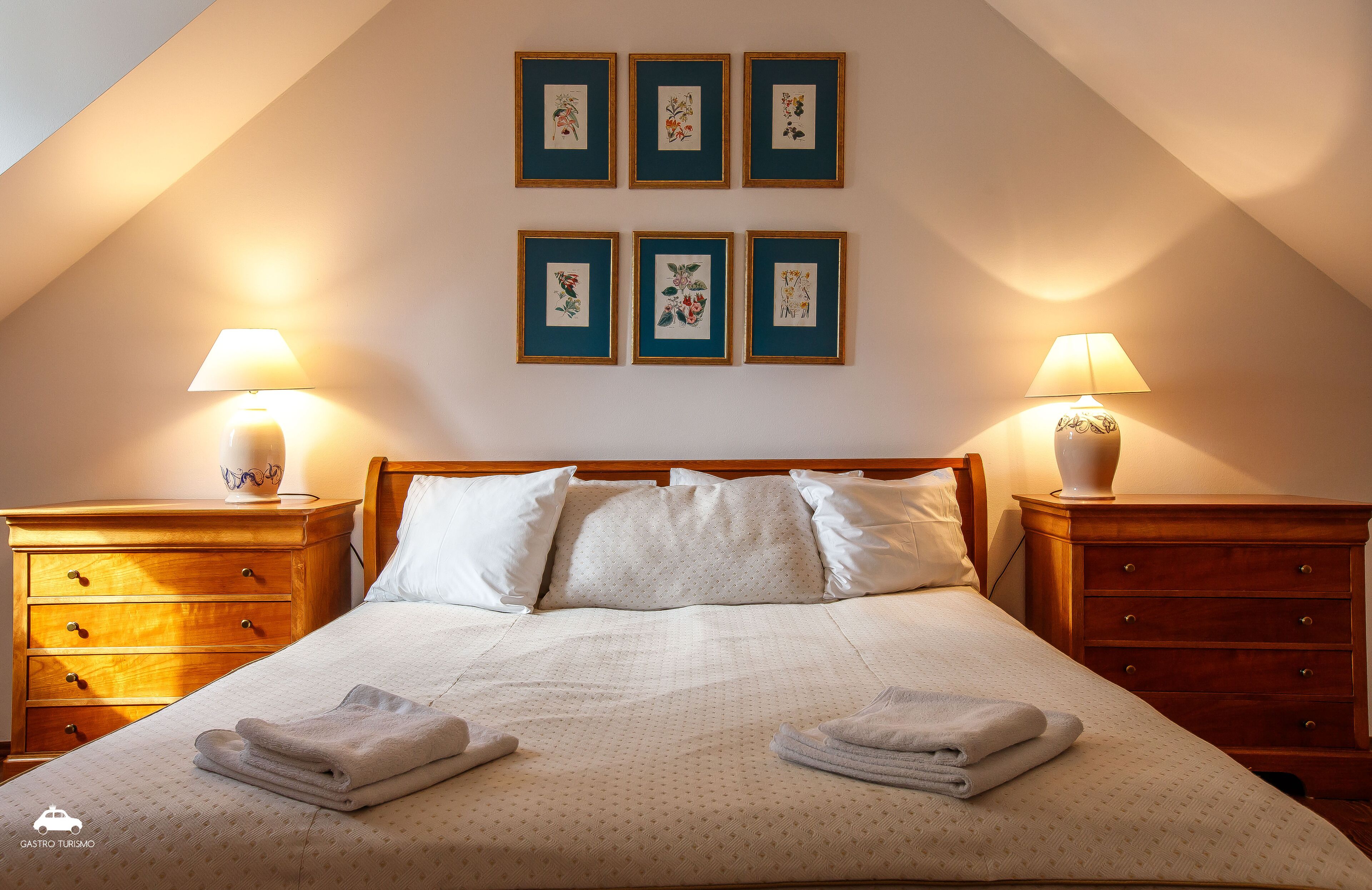 A neatly made bed with a white duvet, two bedside tables with lamps, and six framed botanical prints on the wall.