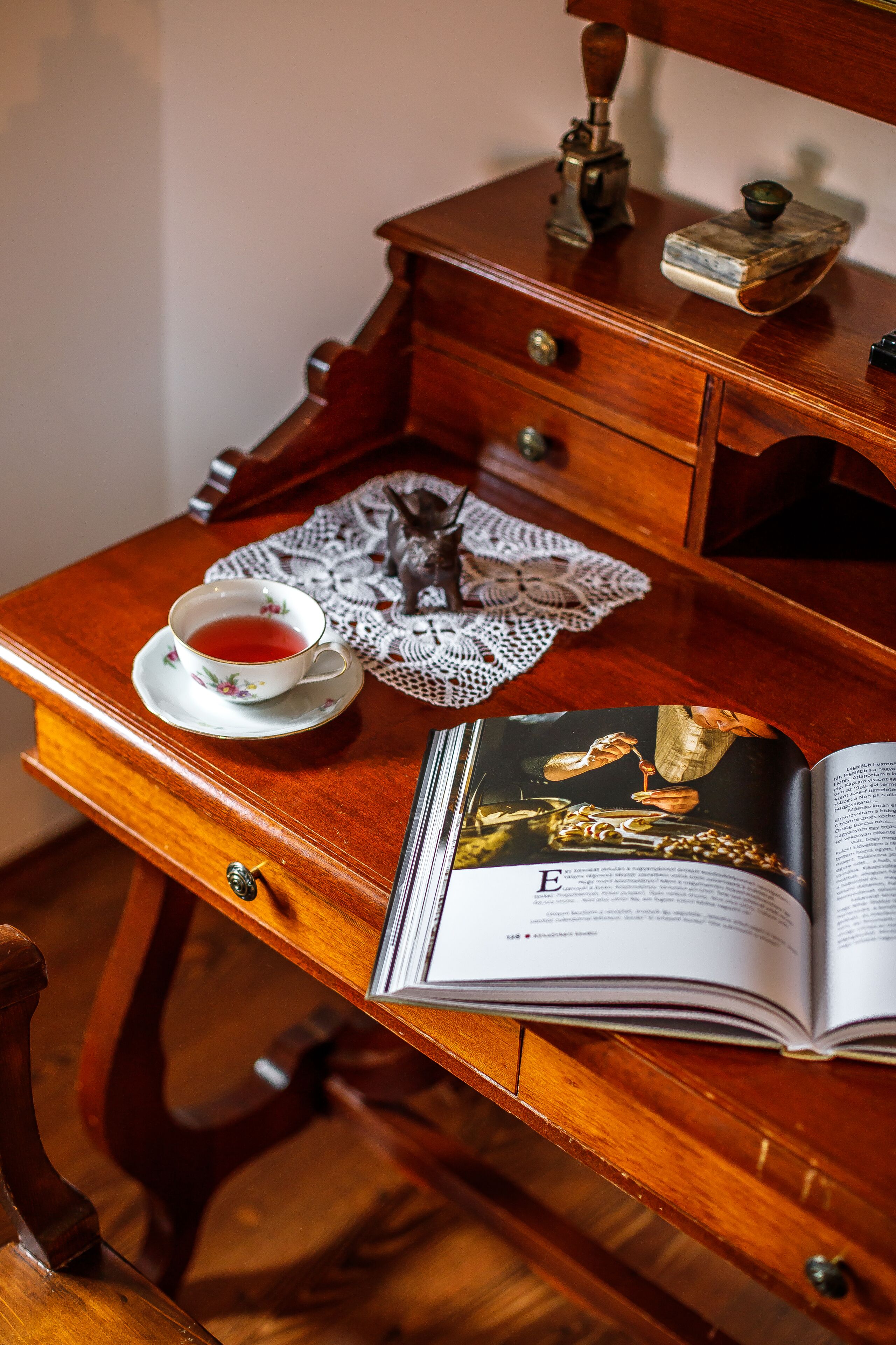 Un escritorio de madera con una taza de té, un posavasos de encaje y un libro abierto a una página que muestra una imagen de joyería.