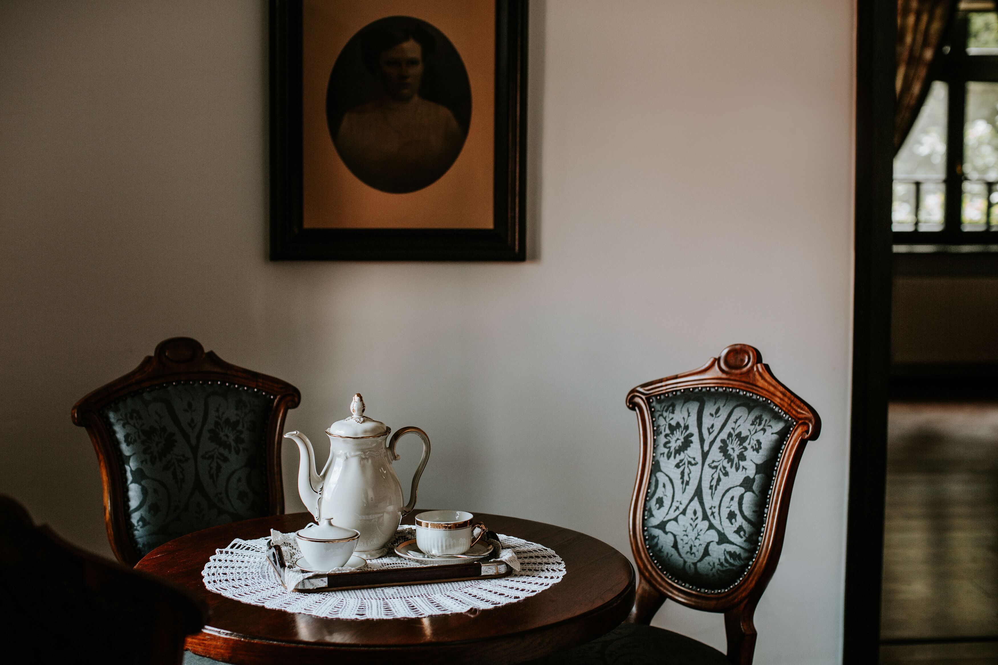 Une table à manger est dressée avec une théière, des tasses et une nappe en dentelle. Deux chaises à motifs floraux sont placées autour de la table. Un portrait encadré est accroché au mur, au-dessus de la table.