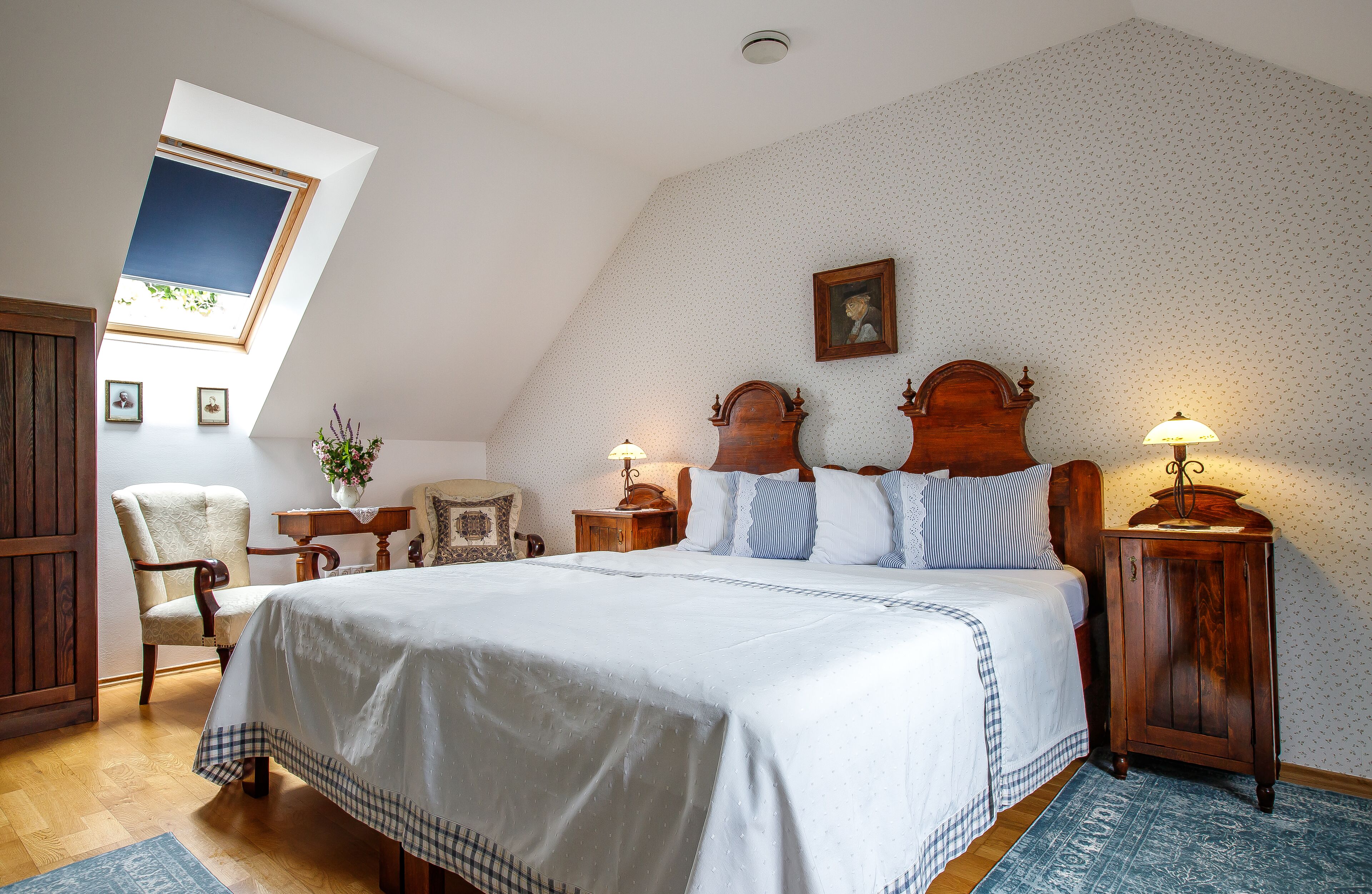 A bedroom with a large bed, wooden headboard, two bedside tables with lamps, a chair, a small table with a vase, and a picture on the wall.