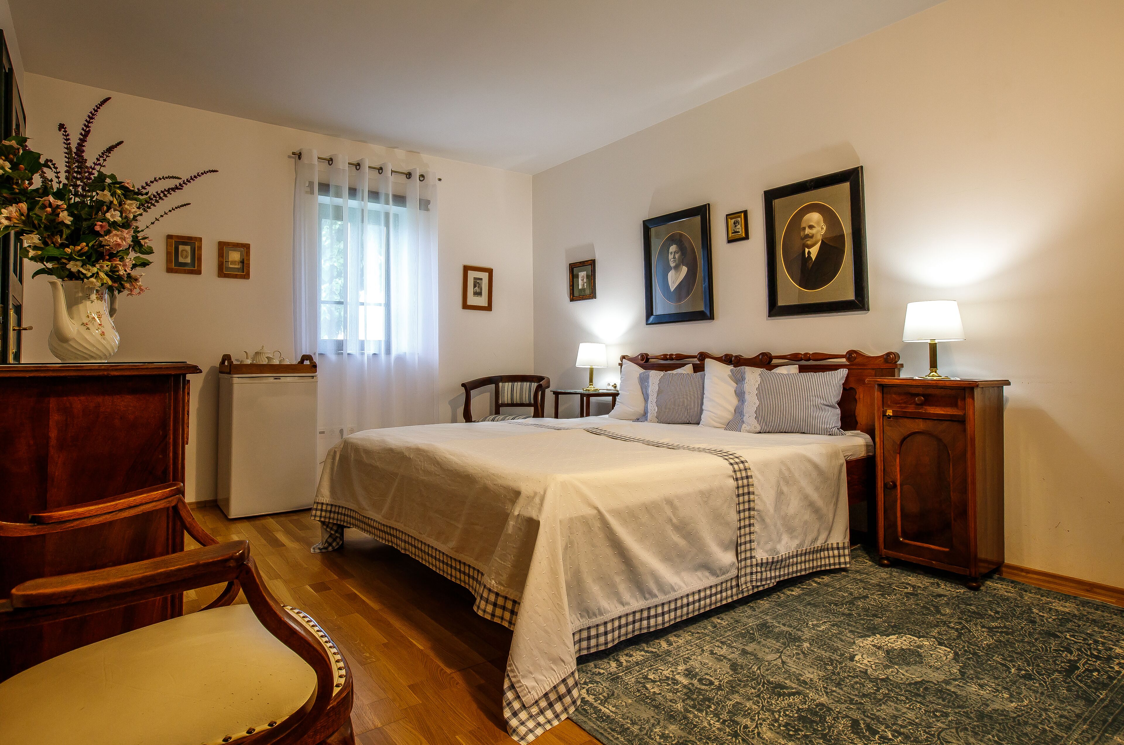 Un dormitorio con cama, mesita de noche, lámpara, silla, escritorio, cómoda, una pequeña mesa, un jarrón con flores y una ventana con cortinas.