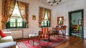 Una habitación con piso de madera, un tapete rojo estampado, una silla, una mesa con lámpara y una ventana con contraventanas verdes.