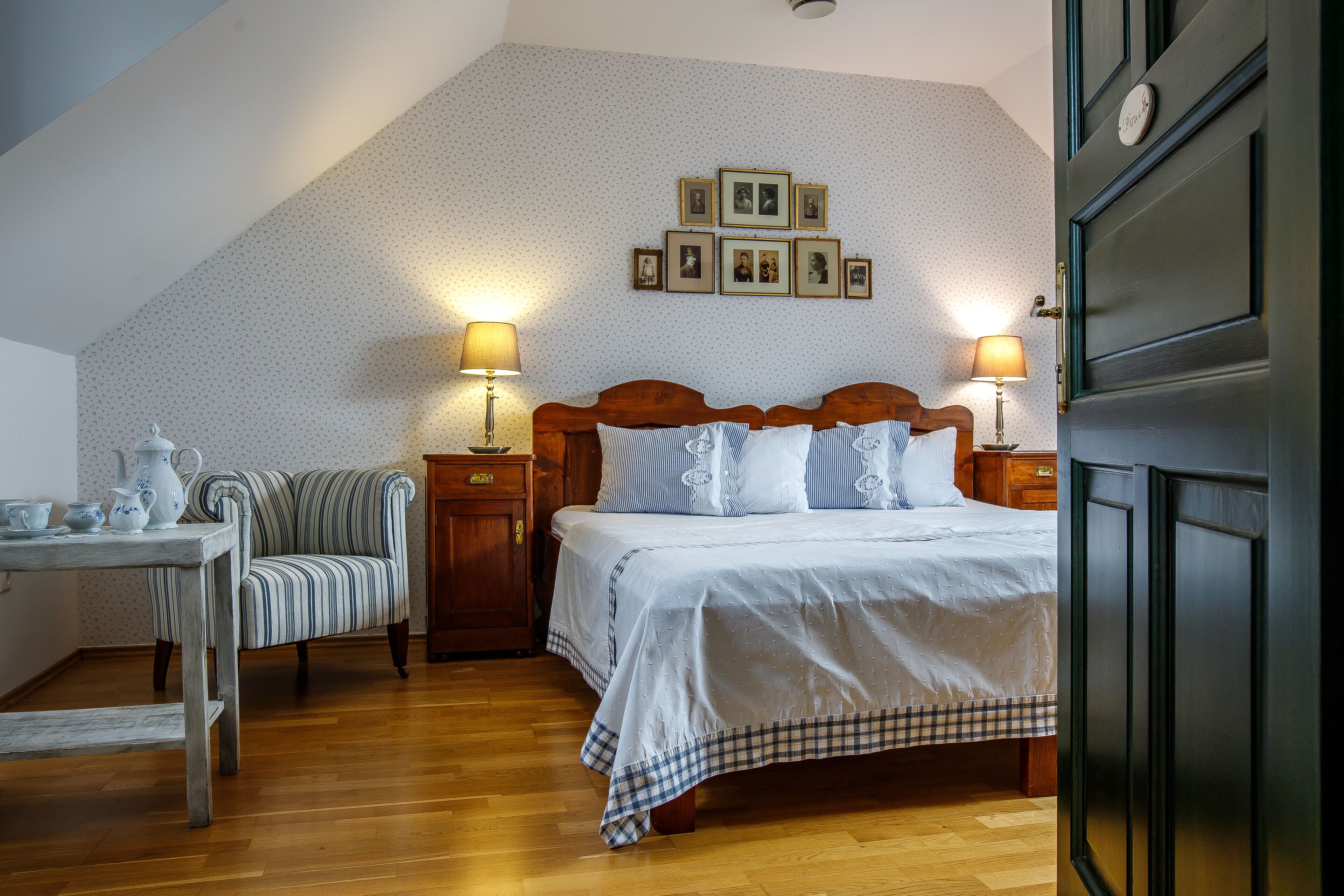 A bedroom with a wooden bed, a chair, a table with teapots, and wall-mounted picture frames.