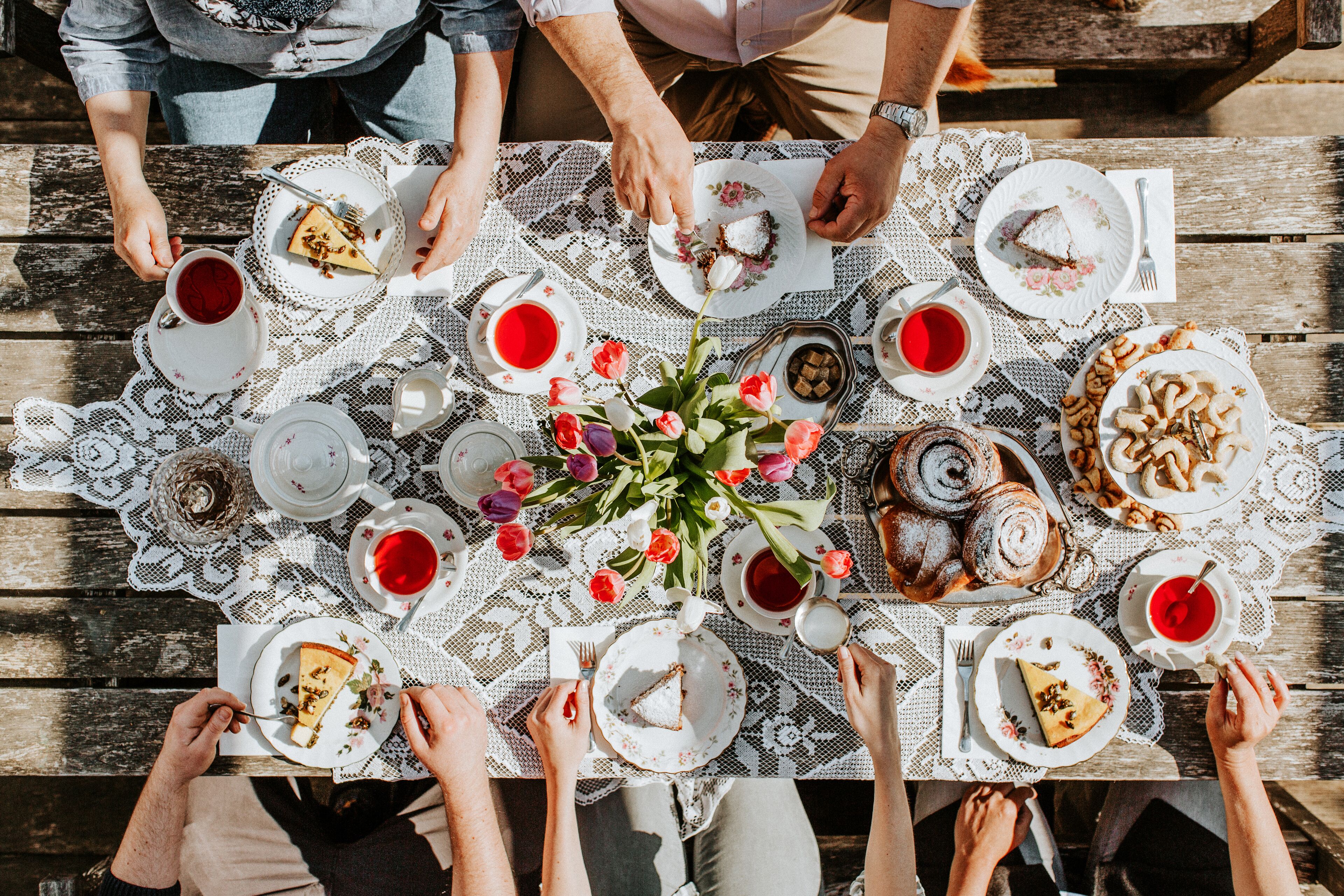 Un grupo de personas sentadas alrededor de una mesa de madera con mantel de encaje, disfrutando de diversos postres y bebidas.