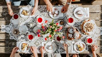 Un grupo de personas sentadas alrededor de una mesa de madera con mantel de encaje, disfrutando de diversos postres y bebidas.