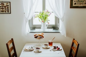 Una mesa servida con pasteles, té y un arreglo floral junto a una ventana con cortinas translúcidas.