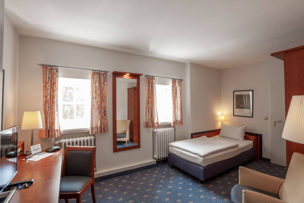 Hotellihuone, jossa on sänky, työpöytä tietokoneella, tuoli, patteri ja ikkuna, jossa on verhot.