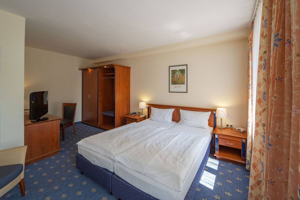Hotellihuone, jossa on suuri sänky, työpöytä, jossa on televisio, vaatekaappi ja yöpöytä, jossa on lamppu.