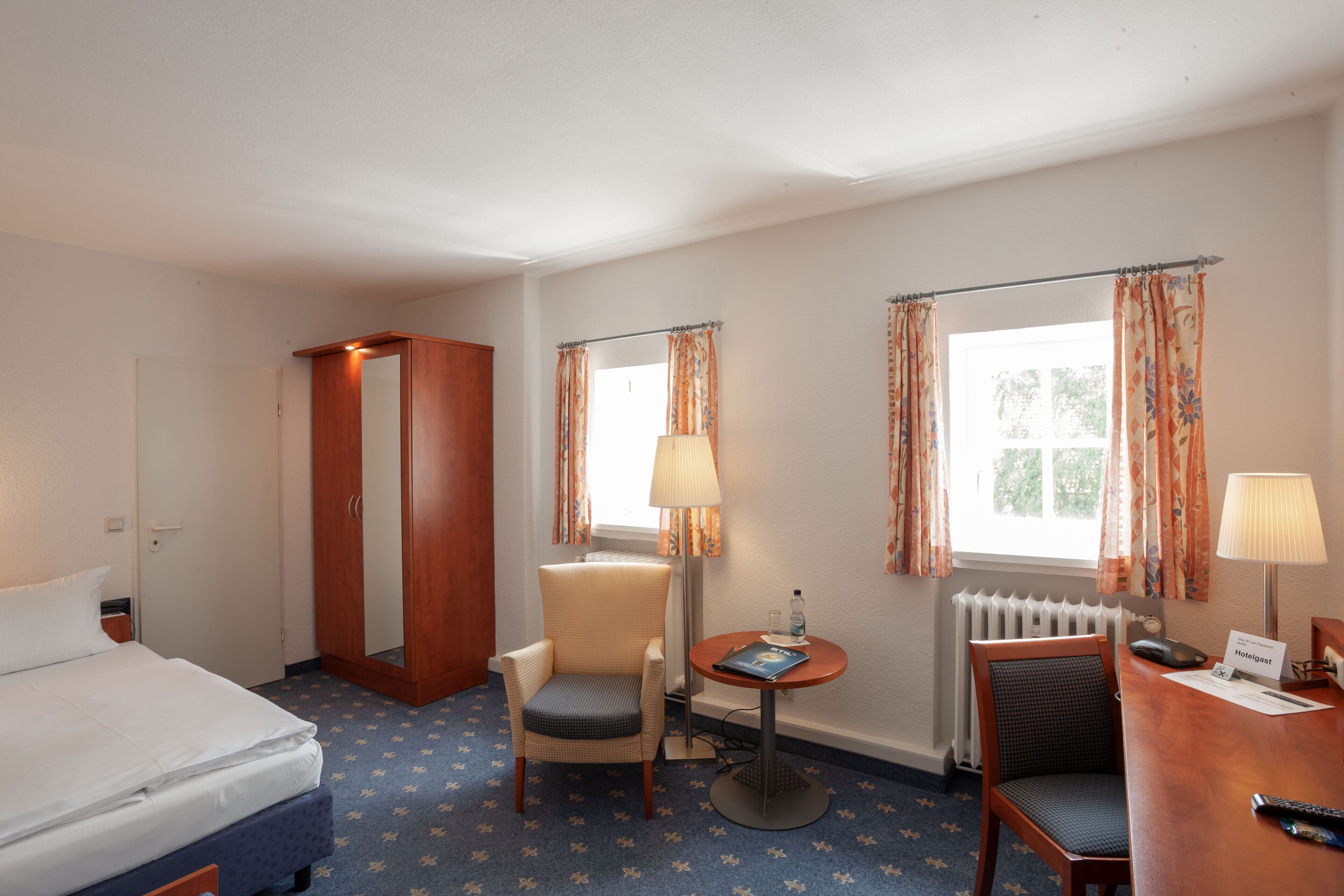 Una habitación de hotel con una cama, dos sillas, una mesita con una lámpara, un radiador y una ventana con cortinas.
