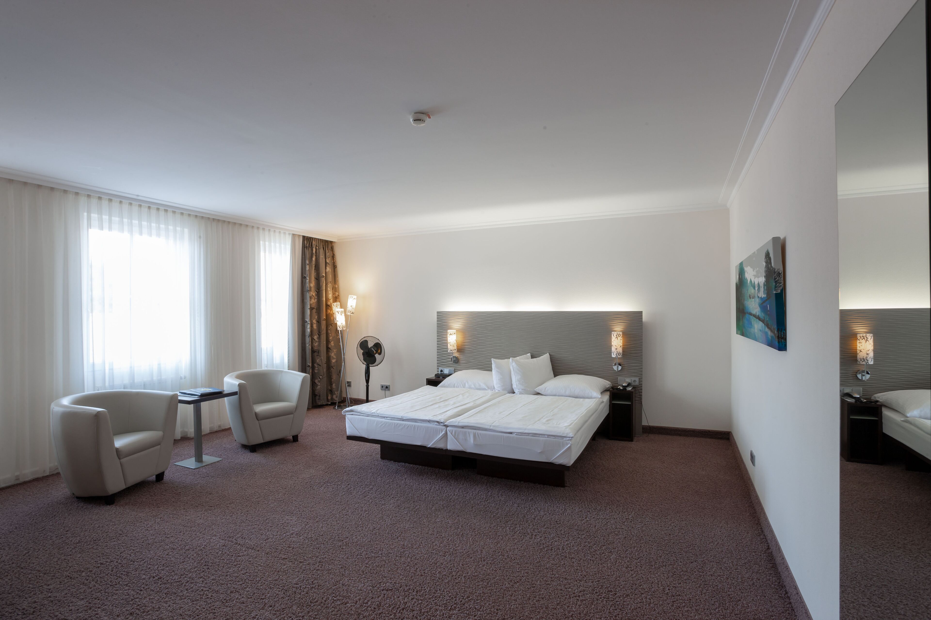 A hotel room with a large bed, two armchairs, a small table, and a nightstand with a lamp.