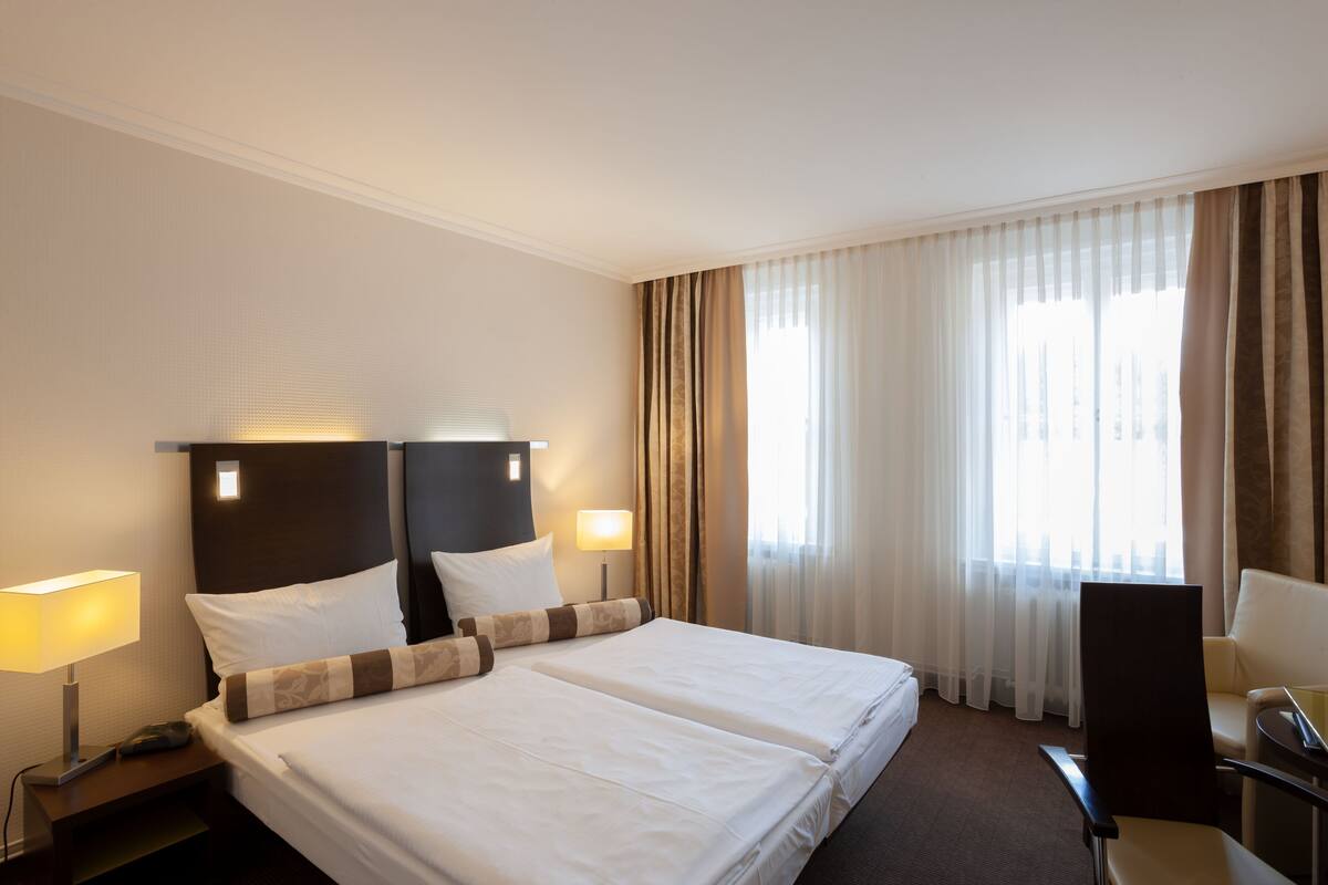 Hotellihuone, jossa on parivuode, kaksi yöpöytävalaisinta, työpöytä tuolilla sekä suuri ikkuna, jossa on verhot.