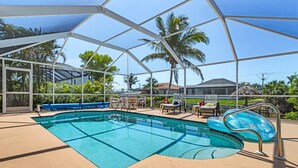 Ein überdachter Swimmingpool mit Liegestühlen und einem Tisch, umgeben von Palmen und blauem Himmel.
