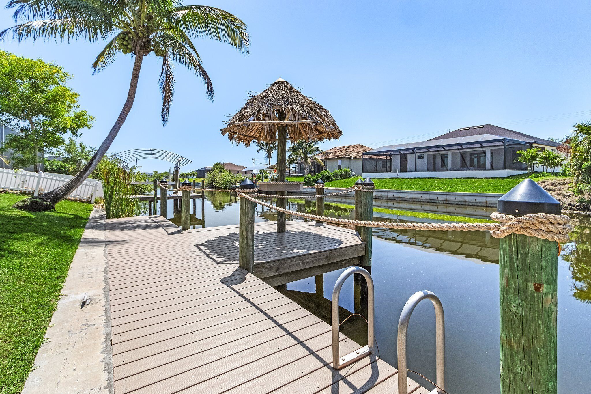 A wooden dock leading to a water body with a thatched-roof structure, surrounded by greenery and a house in the background.