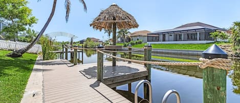 A wooden dock leading to a water body with a thatched-roof structure, surrounded by greenery and a house in the background.