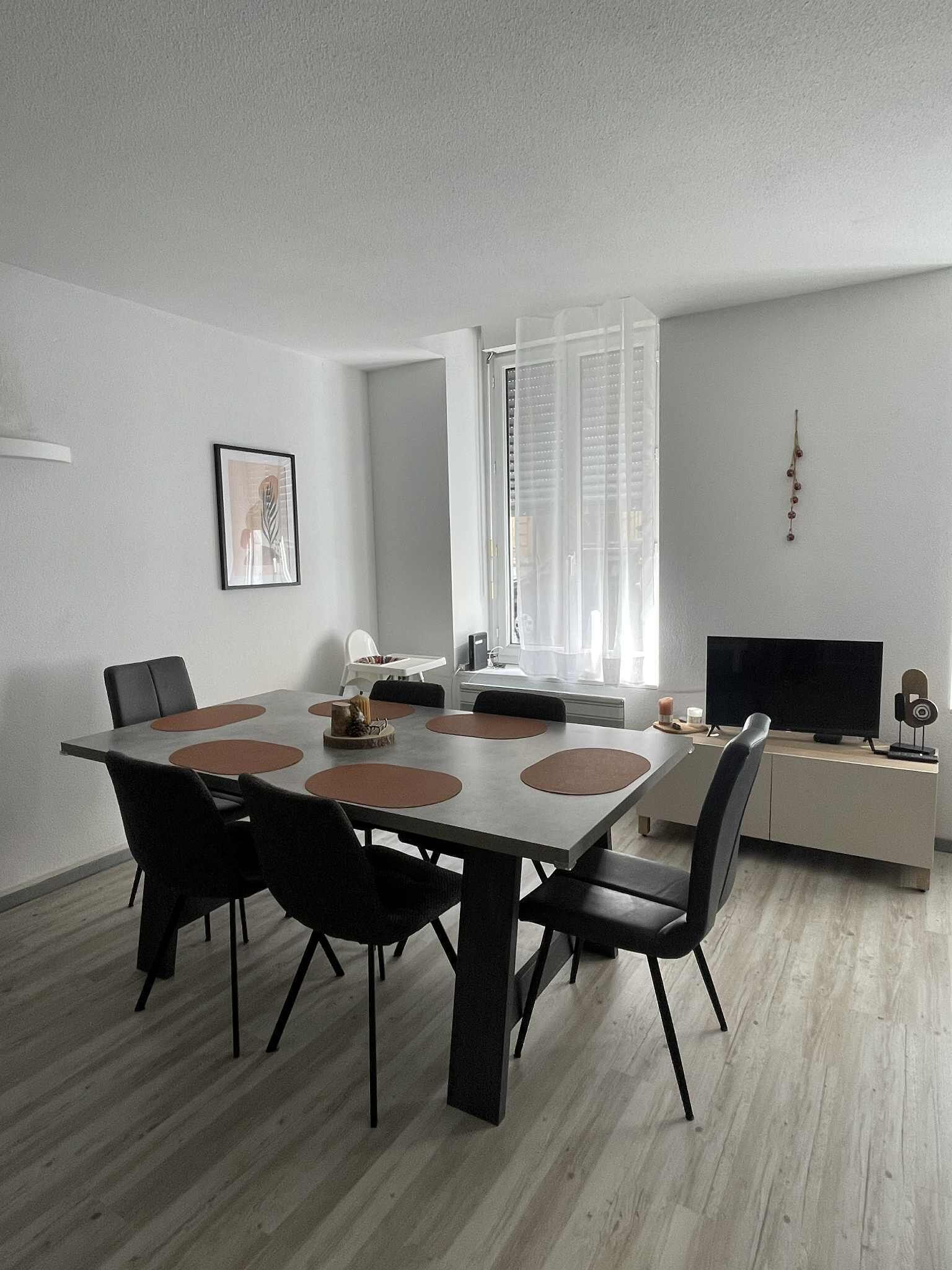 A modern dining area with a large table, chairs, a television, and a window with blinds.