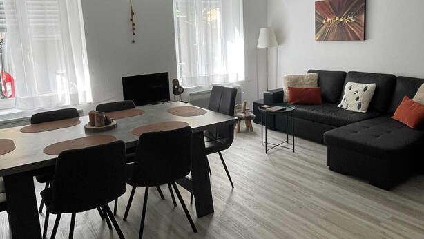 Una sala de estar moderna con comedor, que incluye un sofá negro, una mesa de comedor con sillas y un ventanal con cortinas traslúcidas.