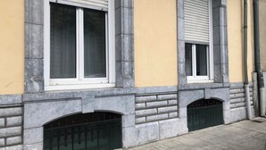 Fachada de edificio con tres ventanas, cada una con marco blanco y persiana gris. Abajo, hay dos ventanas pequeñas y arqueadas, y se ve una valla metálica en frente del edificio.