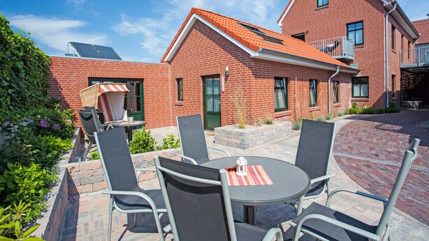 A patio with a round table and four chairs, a brick building with a red roof, and a green door. - Wohnung Bucki (Borkum)