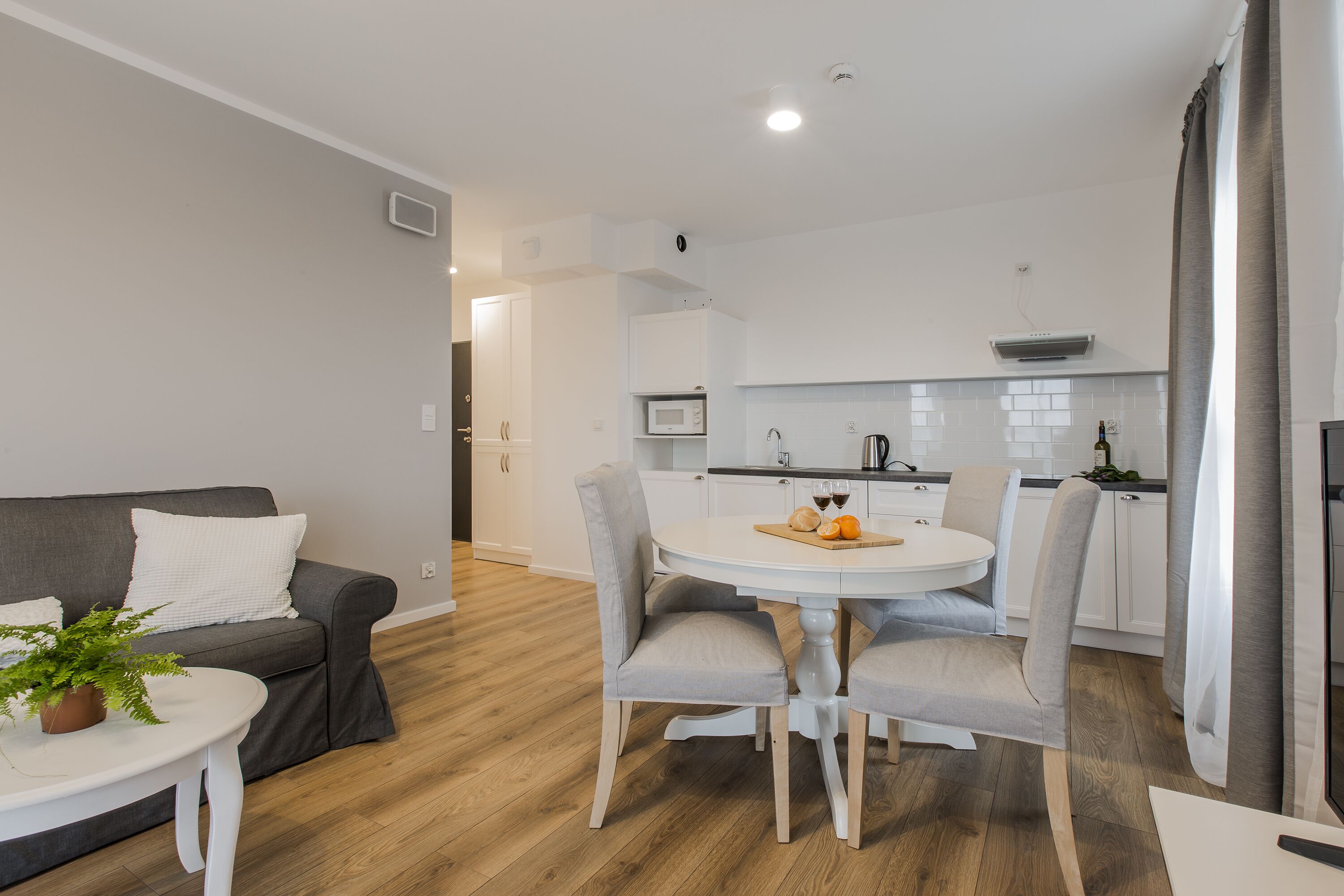 A modern kitchen with white cabinets, a microwave, and a kettle. There is a dining table with four chairs and a potted plant on the side.