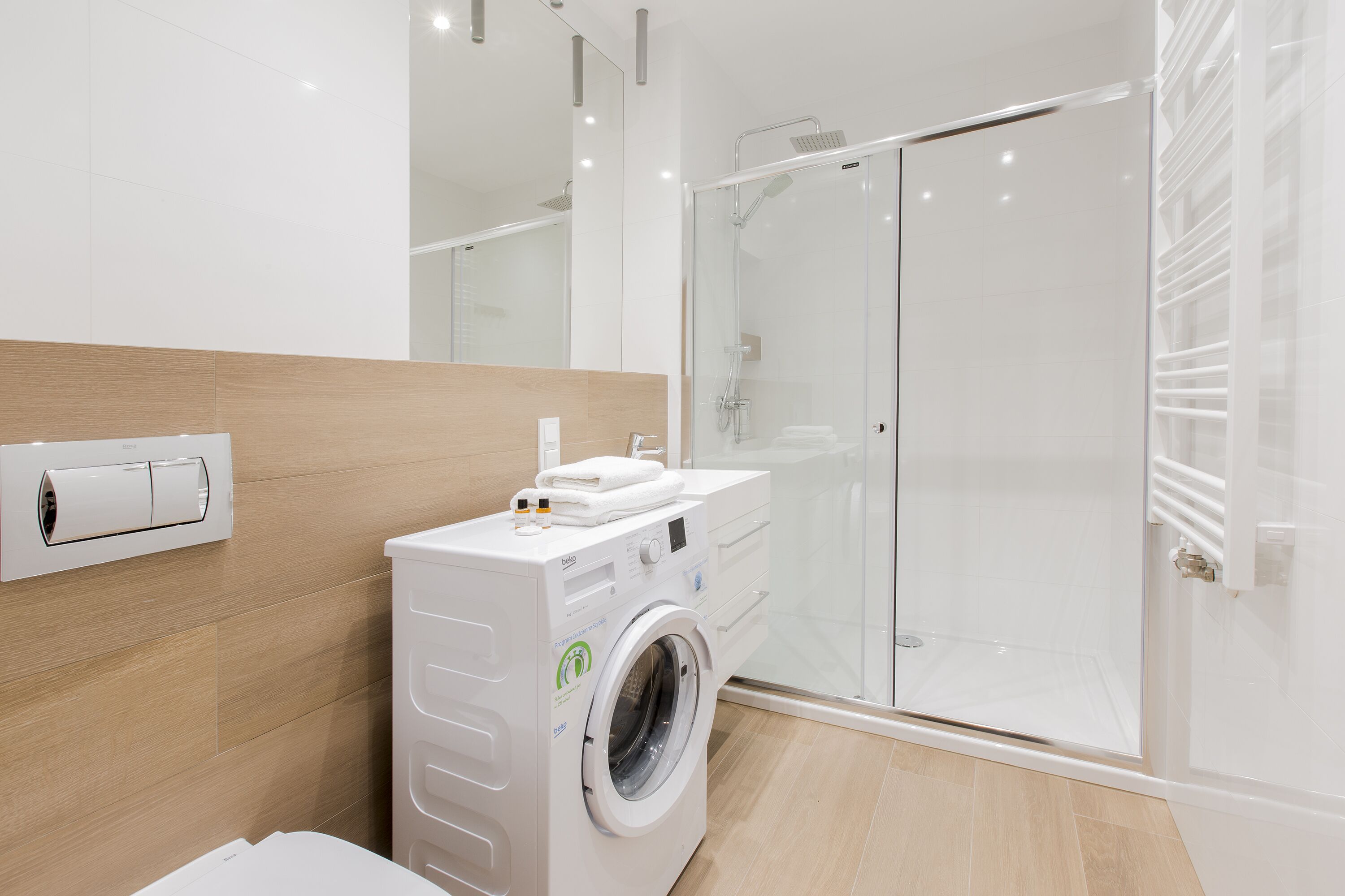A modern bathroom with a built-in washing machine, a glass shower enclosure, and a wooden floor.