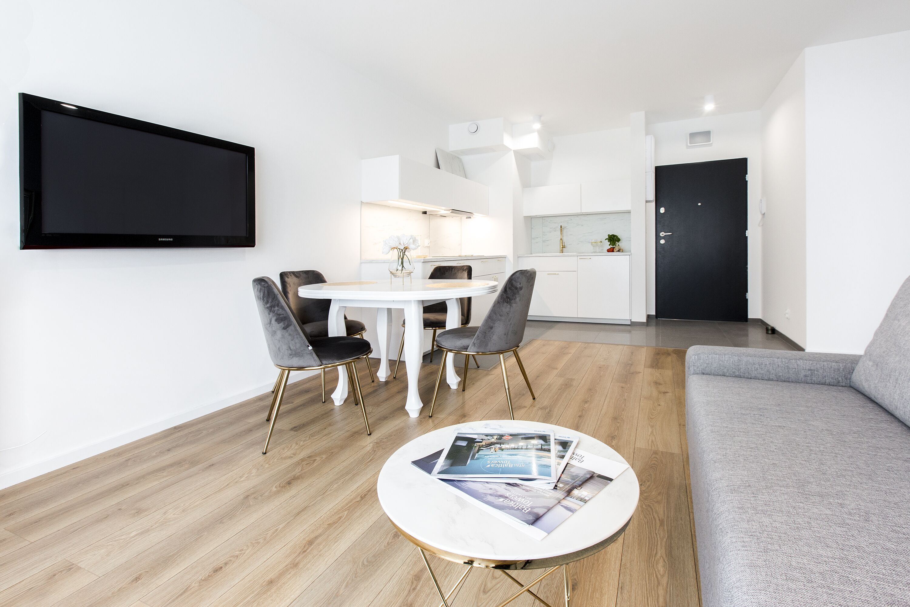 A modern living room with a grey sofa, a round coffee table with magazines, a dining table with four chairs, and a kitchen area in the background.