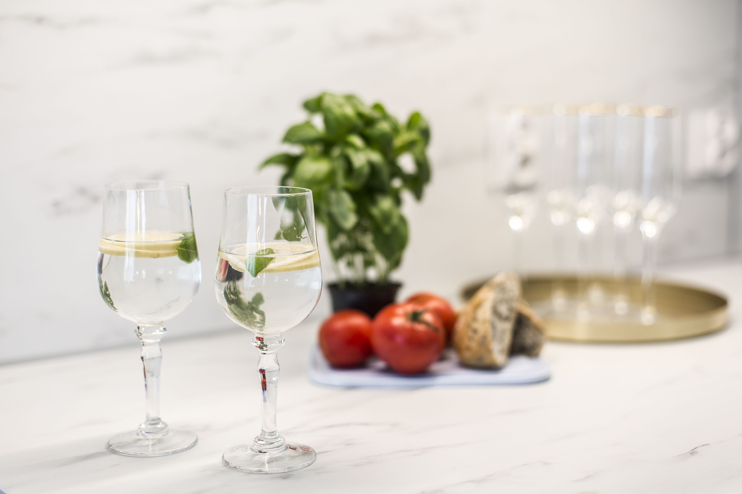 Two glasses of white wine on a marble surface with a potted plant and tomatoes in the background.
