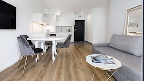 Uma sala de estar moderna com um sofá cinzento, uma mesa de centro redonda e duas cadeiras cinzentas com pés dourados. Uma mesa de jantar branca com uma cadeira branca está ao fundo.