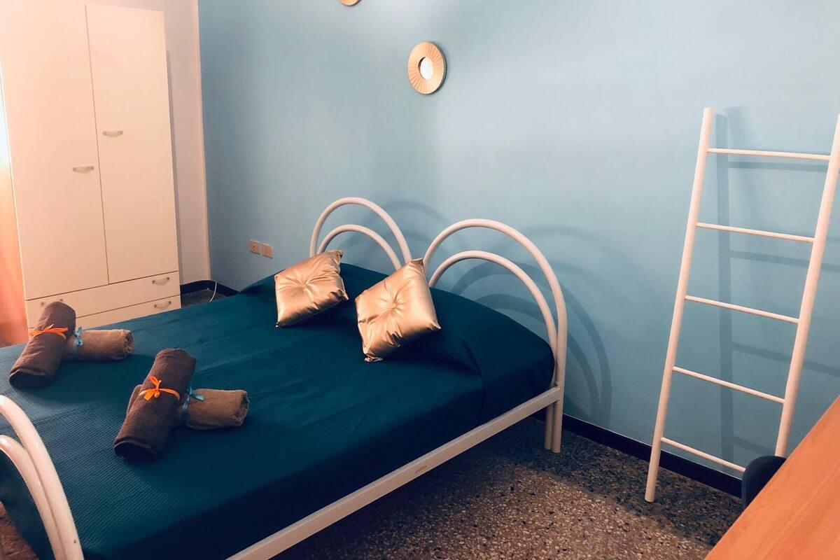 A single bed with a green bedspread, two pillows, and two rolled towels on the bed. There is a white wardrobe to the left and a white ladder to the right.