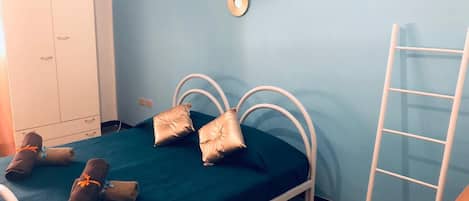 A single bed with a green bedspread, two pillows, and two rolled towels on the bed. There is a white wardrobe to the left and a white ladder to the right.