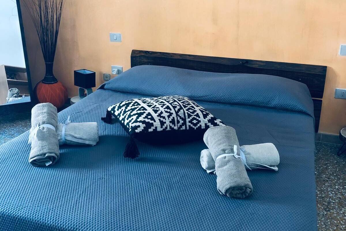 A neatly made bed with blue bedding, a patterned pillow, and rolled towels on a textured blue mat.