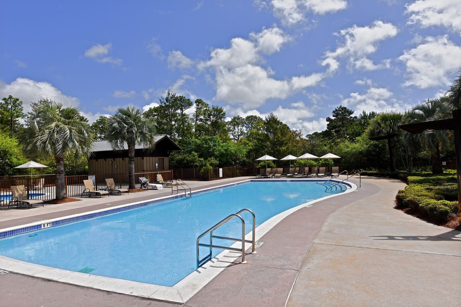 A swimming pool with lounge chairs and umbrellas, surrounded by trees and a clear sky.