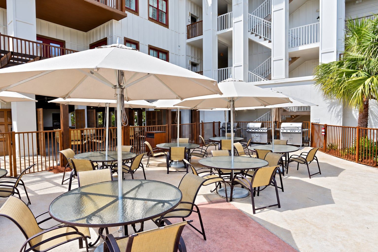 Ein Terrassenbereich im Freien mit runden Tischen und Stühlen, Sonnenschirmen und einem Grill.