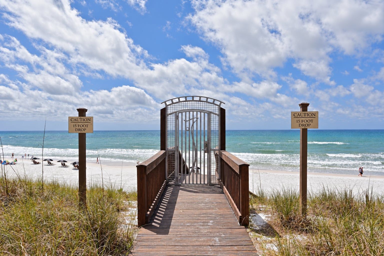 A wooden boardwalk leading to a gated entrance with caution signs indicating a 15-foot drop.